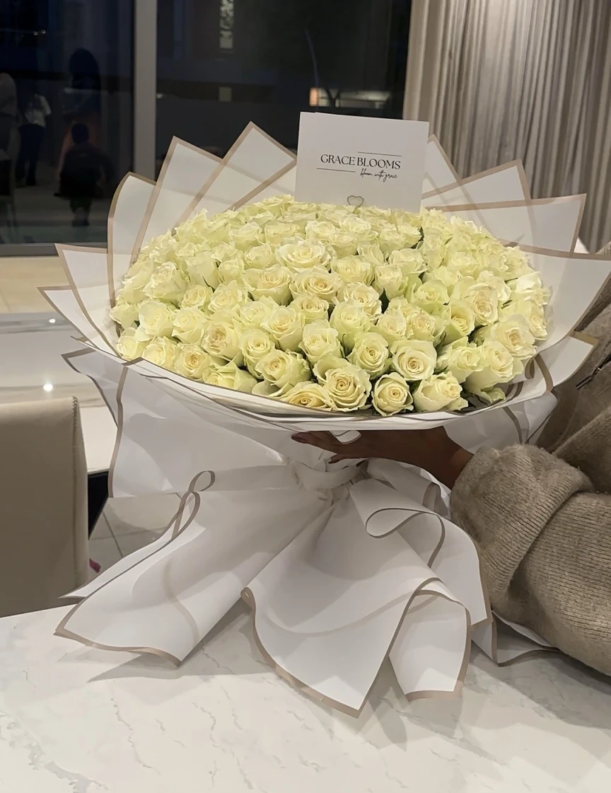 A front-facing ivory rose bouquet in sculptural white and gold-edged wrapping, with a Grace Blooms card